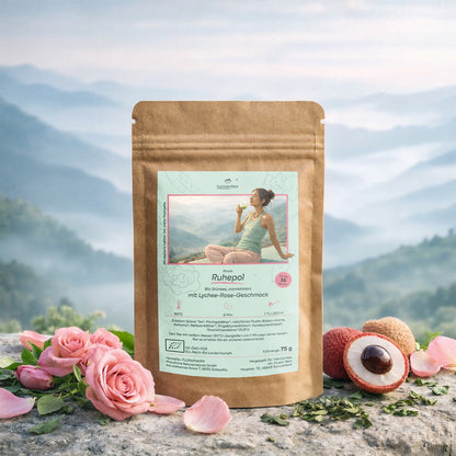 Eine Verpackung von Rosas Ruhepol, dem Bio Grüntee mit Lychee Rose Geschmack auf einem Stein umgeben von Rosenblätter und Litschis